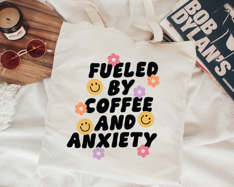 Pode incluir: Sacola de lona branca com o texto "Fueled by coffee and anxiety" em preto, com contorno branco, em uma fonte retr&ocirc;. O texto &eacute; cercado por pequenos rostos sorridentes amarelos e flores rosa e roxas.