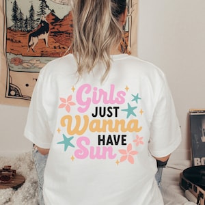 May include: White t-shirt with the text "Girls Just Wanna Have Sun" in pink, yellow, and teal. The design includes floral and star graphics. The shirt is worn by a person with blonde hair.