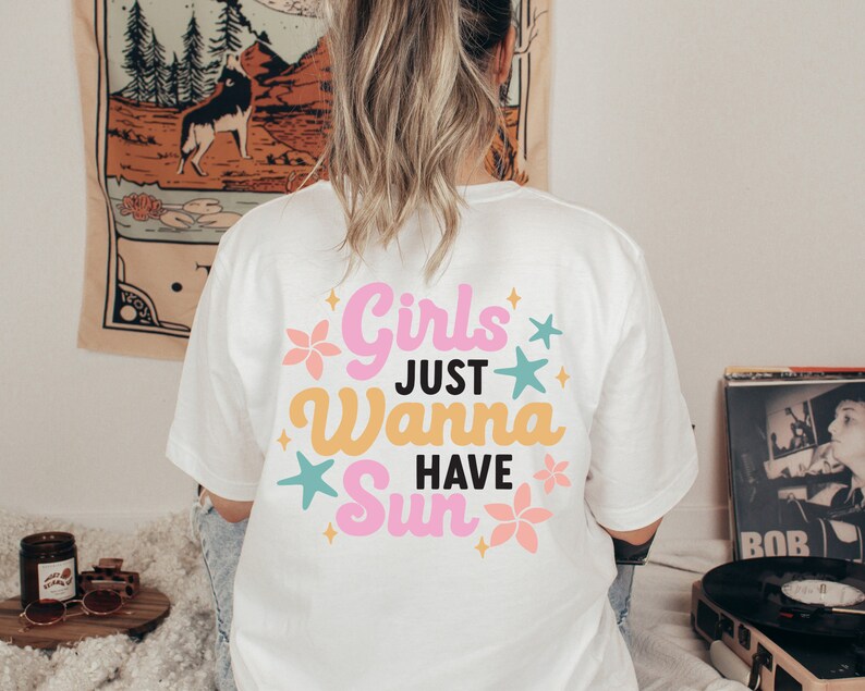 May include: White t-shirt with the text "Girls Just Wanna Have Sun" in pink, yellow, and teal. The design includes floral and star graphics. The shirt is worn by a person with blonde hair.