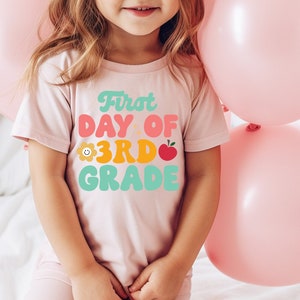 May include: A pink t-shirt with the text "First Day of 3rd Grade" in colorful retro-style lettering. The shirt features a small flower and apple graphic.