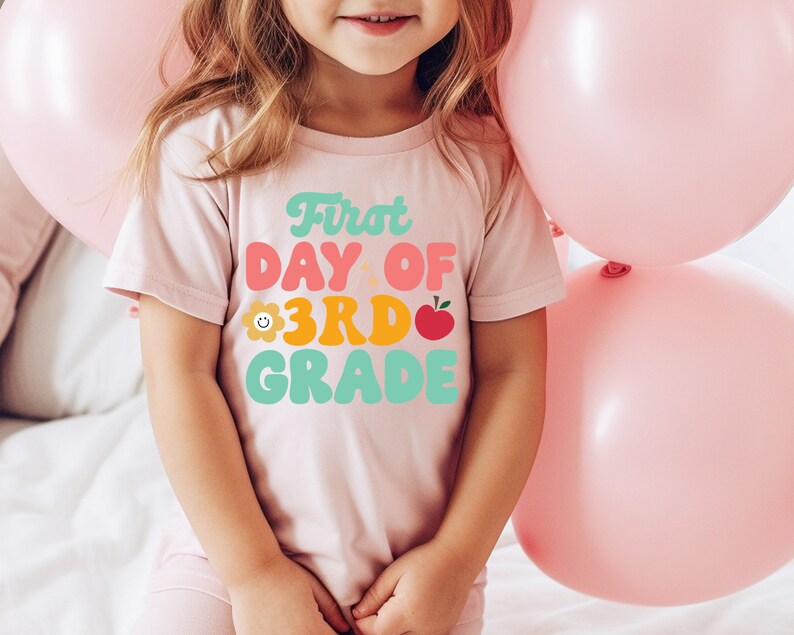 May include: A pink t-shirt with the text "First Day of 3rd Grade" in colorful retro-style lettering. The shirt features a small flower and apple graphic.