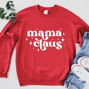 May include: Red sweatshirt with white text that says "mama claus!" with white stars around the text. The sweatshirt is laying on a white surface with a pair of blue jeans and white sneakers.