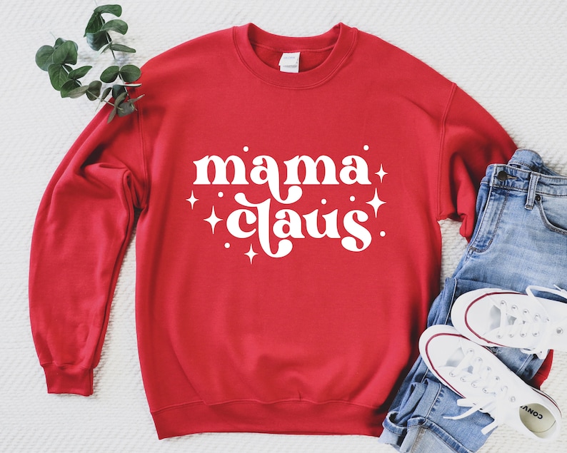May include: Red sweatshirt with white text that says "mama claus!" with white stars around the text. The sweatshirt is laying on a white surface with a pair of blue jeans and white sneakers.