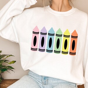 May include: White sweatshirt featuring a colorful crayon design. The design includes six crayons in pink, purple, blue, green, yellow, and orange, each with a black oval and black stripes.
