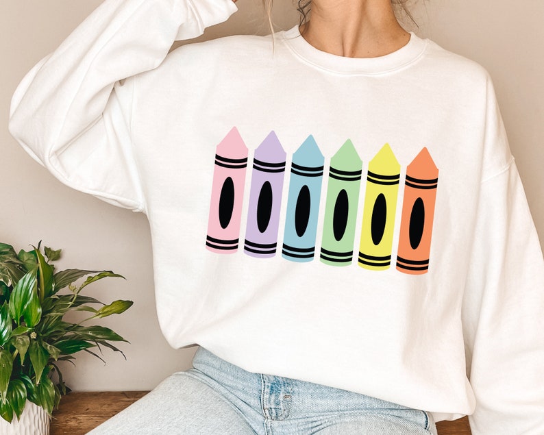 May include: White sweatshirt featuring a colorful crayon design. The design includes six crayons in pink, purple, blue, green, yellow, and orange, each with a black oval and black stripes.