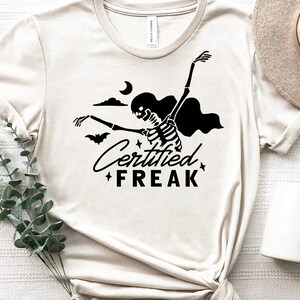May include: A white t-shirt with a black skeleton graphic and the text "Certified Freak" printed on it. The skeleton is wearing a cape and holding a bat. There is a crescent moon and clouds in the background.
