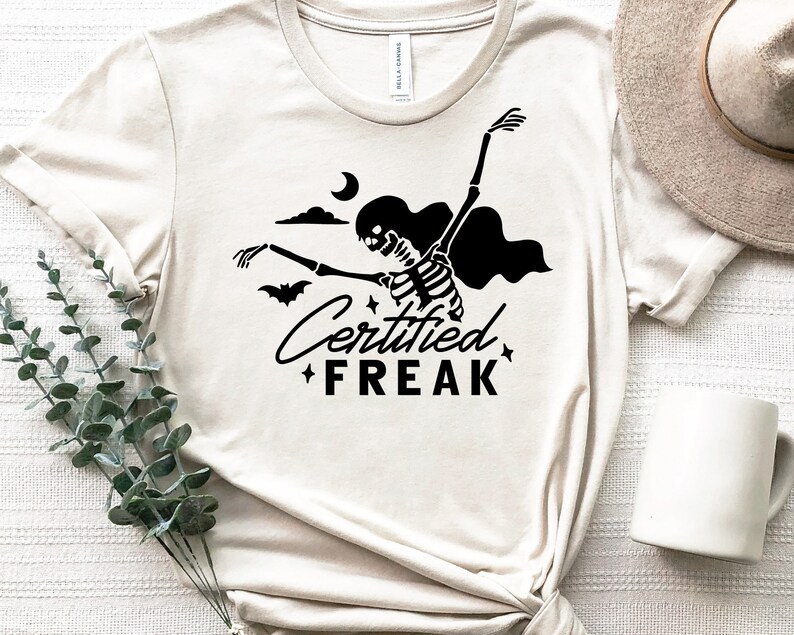 May include: A white t-shirt with a black skeleton graphic and the text "Certified Freak" printed on it. The skeleton is wearing a cape and holding a bat. There is a crescent moon and clouds in the background.