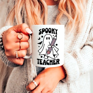 Peut inclure: Une tasse en c&eacute;ramique blanche avec une illustration en noir et blanc d'un fant&ocirc;me tenant un crayon. Le texte "Spooky Teacher" est imprim&eacute; en noir sur la tasse.
