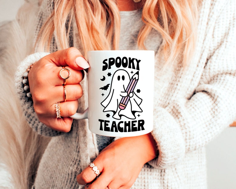 Peut inclure: Une tasse en c&eacute;ramique blanche avec une illustration en noir et blanc d'un fant&ocirc;me tenant un crayon. Le texte "Spooky Teacher" est imprim&eacute; en noir sur la tasse.