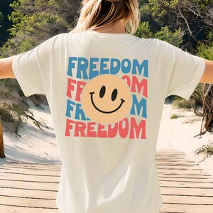 May include: White t-shirt featuring a graphic of a smiley face and the word "FREEDOM" repeated in a layered design with blue, red, and pink colours.