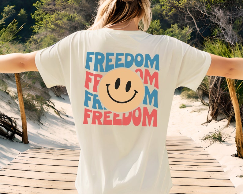 May include: White t-shirt featuring a graphic of a smiley face and the word "FREEDOM" repeated in a layered design with blue, red, and pink colours.