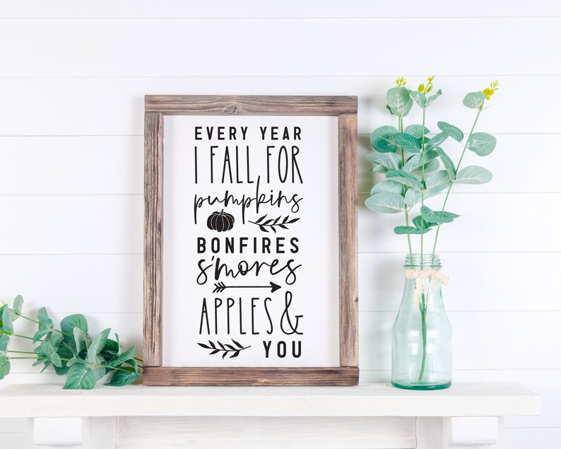 K&ouml;nnte beinhalten: Ein rustikales Holzschild mit dem Text "Every year I fall for pumpkins, bonfires, s'mores, apples & you" in schwarzer Schrift. Das Schild ist mit einer kleinen K&uuml;rbisgrafik, einem kleinen Pfeil und einem kleinen Zweig mit Bl&auml;ttern verziert.