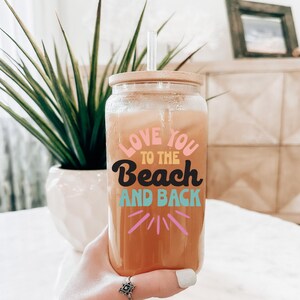 May include: A clear glass tumbler with a bamboo lid and straw, filled with a beverage. The tumbler features the text "LOVE YOU TO THE BEACH AND BACK" in colorful lettering. A green plant in a white pot is in the background.