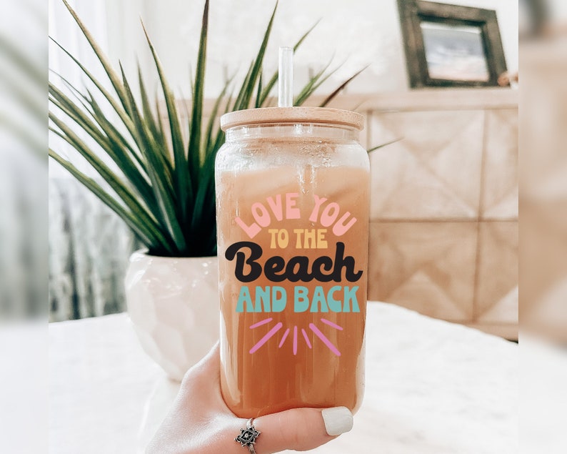 May include: A clear glass tumbler with a bamboo lid and straw, filled with a beverage. The tumbler features the text "LOVE YOU TO THE BEACH AND BACK" in colorful lettering. A green plant in a white pot is in the background.