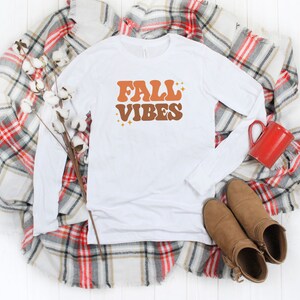 Pode incluir: Camiseta branca de manga comprida com o texto "FALL VIBES" em letras laranja e marrom com detalhes de glitter dourado. A camiseta est&aacute; colocada sobre um cobertor xadrez com uma caneca vermelha e botas marrons.