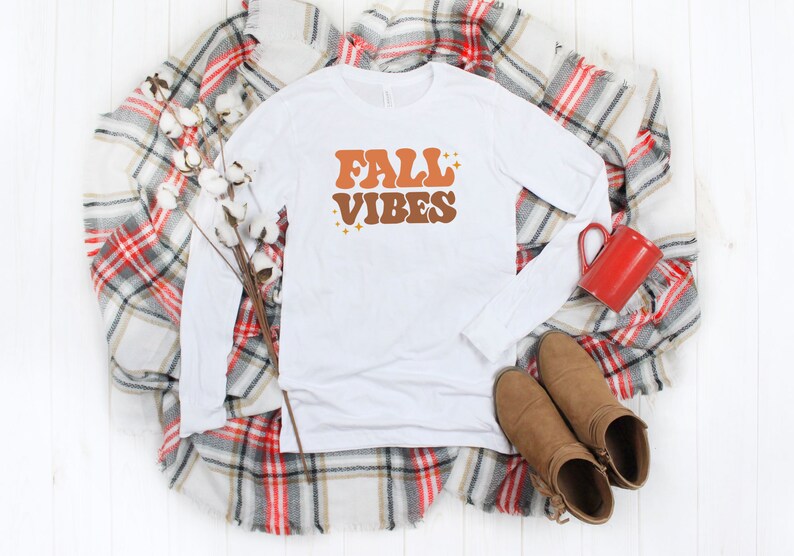 Pode incluir: Camiseta branca de manga comprida com o texto "FALL VIBES" em letras laranja e marrom com detalhes de glitter dourado. A camiseta est&aacute; colocada sobre um cobertor xadrez com uma caneca vermelha e botas marrons.