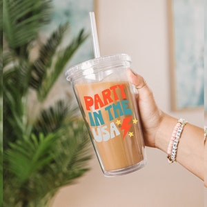 May include: A clear plastic tumbler with a lid and straw. The tumbler has a red, blue and yellow graphic that says "Party in the USA" with stars and a lightning bolt.