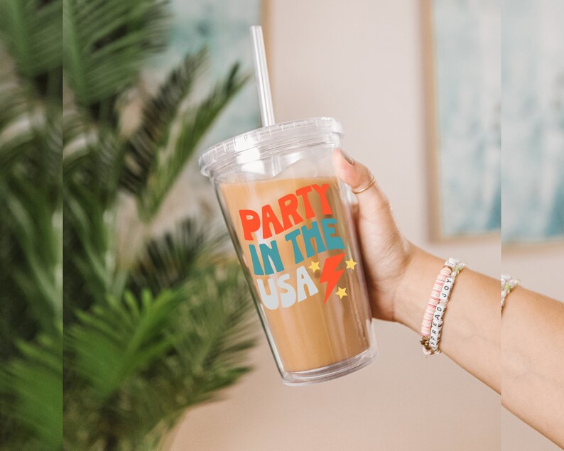 May include: A clear plastic tumbler with a lid and straw. The tumbler has a red, blue and yellow graphic that says "Party in the USA" with stars and a lightning bolt.