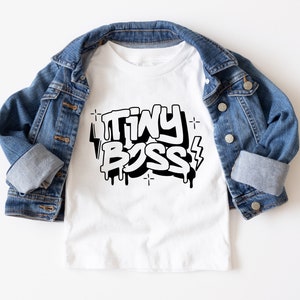 May include: White t-shirt with a black graphic that says "TINY BOSS" in a graffiti style with drips and lightning bolts.