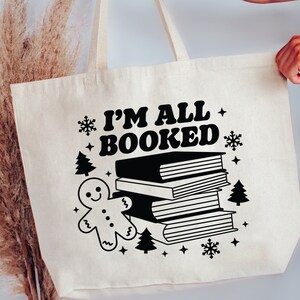 May include: A white canvas tote bag with black text that reads "I'm All Booked" and a black illustration of a gingerbread man and a stack of books. The bag has black snowflake and star accents.