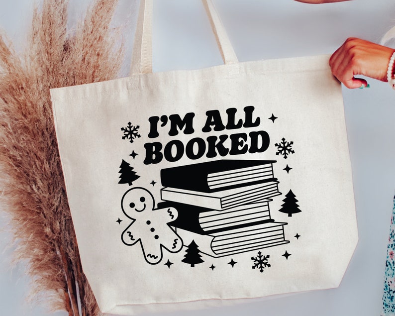 May include: A white canvas tote bag with black text that reads "I'm All Booked" and a black illustration of a gingerbread man and a stack of books. The bag has black snowflake and star accents.