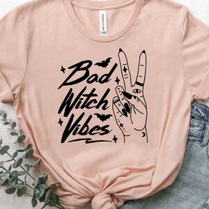 May include: A pink t-shirt with a black graphic design that says "Bad Witch Vibes" with a hand making the peace sign with a crescent moon, stars, and bats.