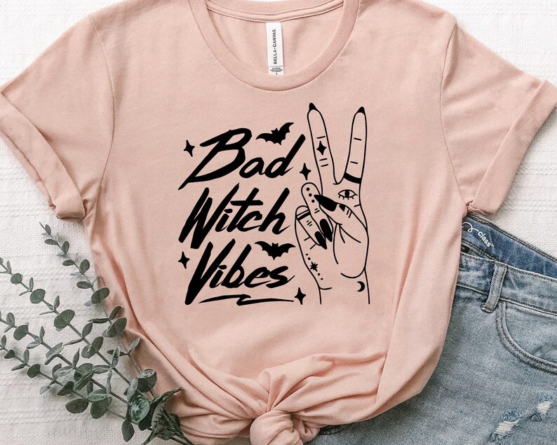 May include: A pink t-shirt with a black graphic design that says "Bad Witch Vibes" with a hand making the peace sign with a crescent moon, stars, and bats.