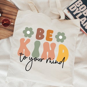 Op de afbeelding: Een witte tote bag met de tekst "Be Kind to your mind" in kleurrijke, retro-stijl letters. De letters zijn zwart omrand en hebben een licht 3D-effect. Twee groene bloemen met witte centra bevinden zich aan weerszijden van de tekst.