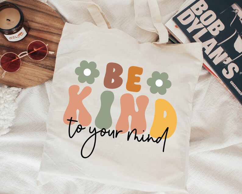 Op de afbeelding: Een witte tote bag met de tekst "Be Kind to your mind" in kleurrijke, retro-stijl letters. De letters zijn zwart omrand en hebben een licht 3D-effect. Twee groene bloemen met witte centra bevinden zich aan weerszijden van de tekst.