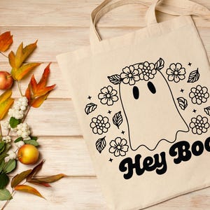 May include: A white tote bag with a black and white illustration of a ghost wearing a flower crown. The text "Hey Boo" is written in a retro font below the ghost.