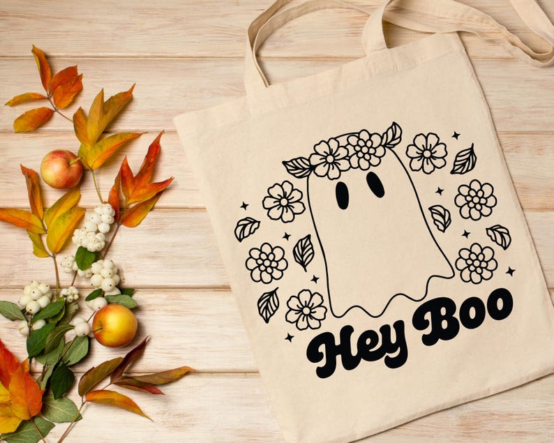 May include: A white tote bag with a black and white illustration of a ghost wearing a flower crown. The text "Hey Boo" is written in a retro font below the ghost.