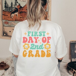 May include: White t-shirt with a colorful graphic that says "First Day of 2nd Grade" with a smiley face flower on either side of the "2nd".