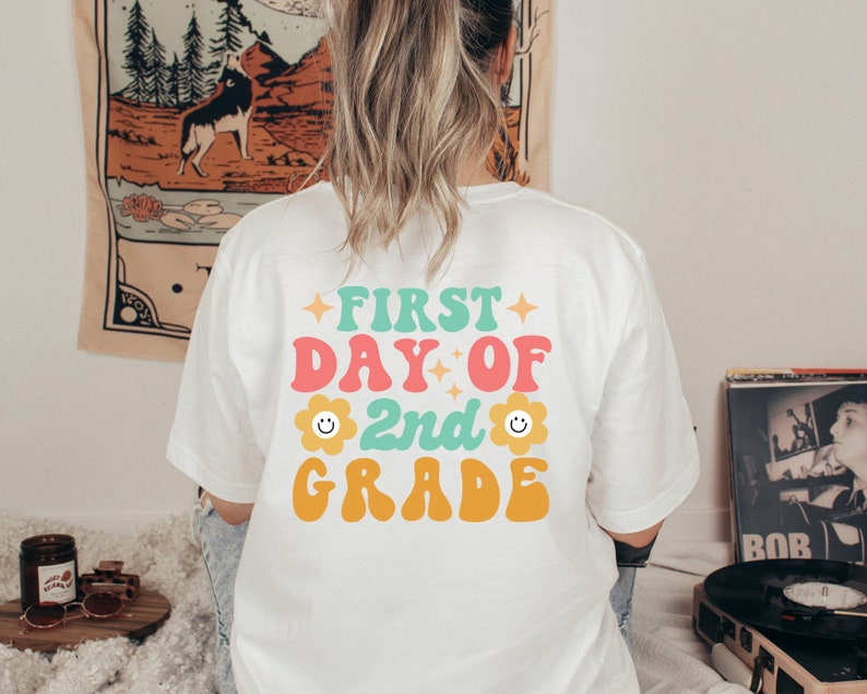 May include: White t-shirt with a colorful graphic that says "First Day of 2nd Grade" with a smiley face flower on either side of the "2nd".