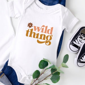 May include: White baby onesie with the text "wild thing" in brown, orange, and pink lettering with a small flower graphic above the text.