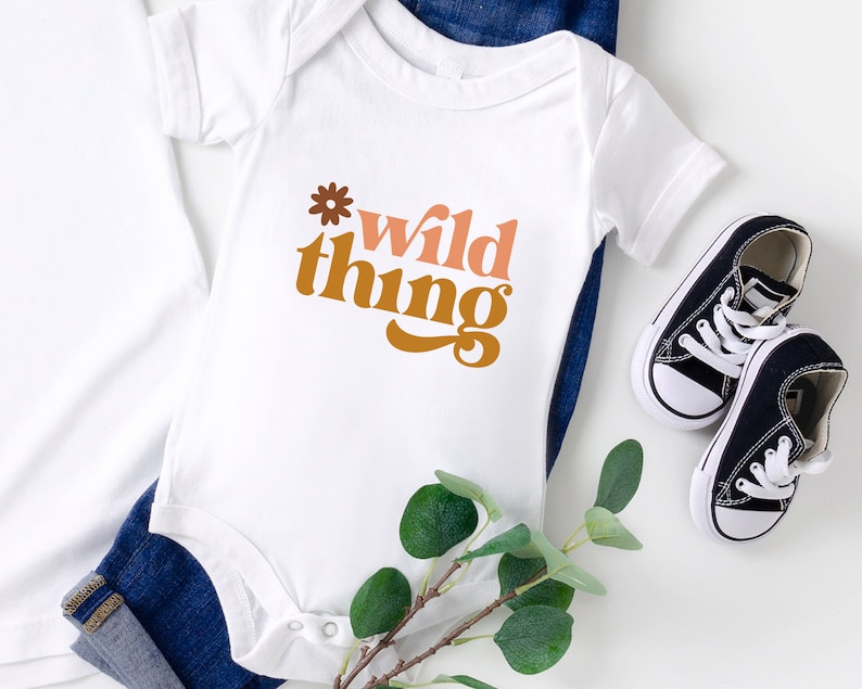 May include: White baby onesie with the text "wild thing" in brown, orange, and pink lettering with a small flower graphic above the text.