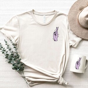 Pu&ograve; includere: Una maglietta beige con una mano viola con le dita incrociate e stelle sul petto sinistro. La maglietta &egrave; appoggiata su una superficie bianca con un cappello e una tazza con lo stesso design.