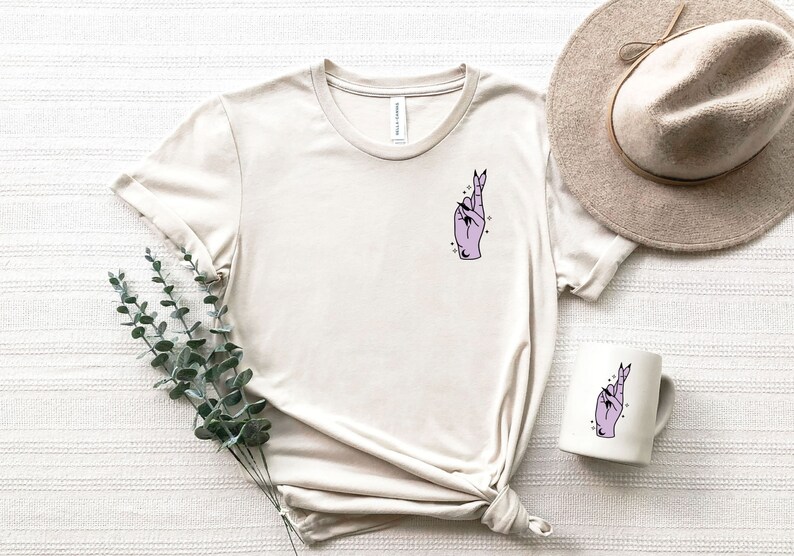 Pu&ograve; includere: Una maglietta beige con una mano viola con le dita incrociate e stelle sul petto sinistro. La maglietta &egrave; appoggiata su una superficie bianca con un cappello e una tazza con lo stesso design.