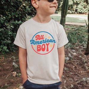 May include: A white t-shirt with a red, blue and yellow circle graphic that says "All American Boy" with stars around the circle.