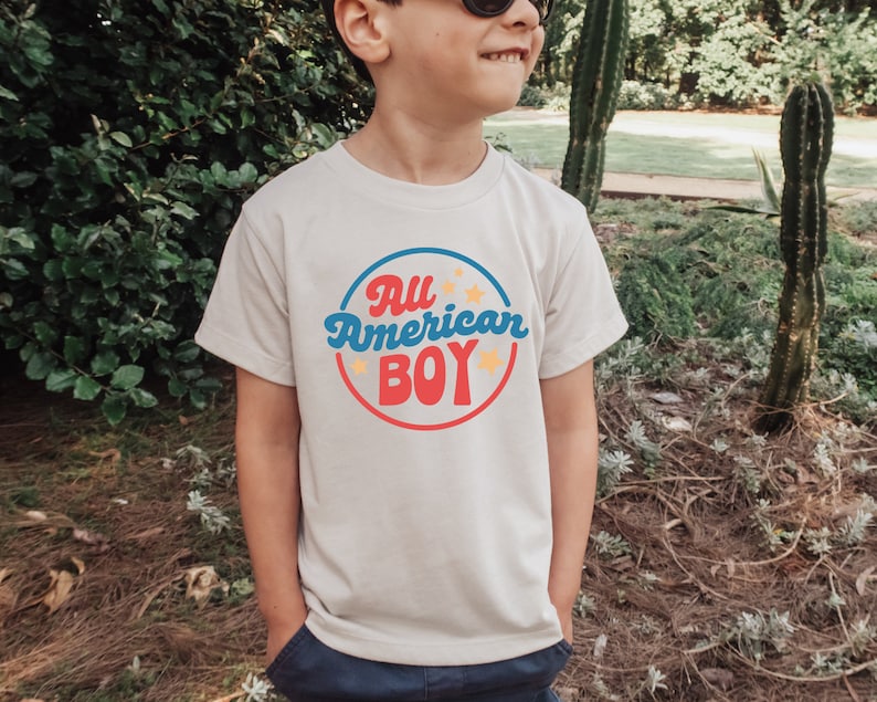 May include: A white t-shirt with a red, blue and yellow circle graphic that says "All American Boy" with stars around the circle.