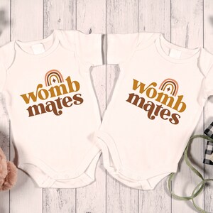 May include: Two white baby onesies with brown text that reads "womb mates" and a rainbow graphic.
