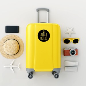 K&ouml;nnte beinhalten: Gelber Koffer mit einem schwarzen runden Aufkleber, auf dem "Aqua Holic" mit einem Anker-Symbol steht. Der Koffer ist von Reiseaccessoires umgeben, darunter ein Strohhut, eine Sonnenbrille, eine Kamera, Sonnencreme und ein Seestern.