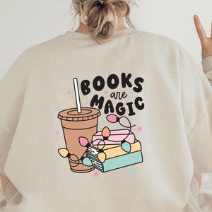 K&ouml;nnte beinhalten: Ein beiger Pullover mit einem grafischen Design aus einer Tasse Kaffee, einem Stapel B&uuml;chern und dem Text "BOOKS are MAGIC".
