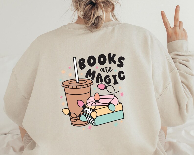 K&ouml;nnte beinhalten: Ein beiger Pullover mit einem grafischen Design aus einer Tasse Kaffee, einem Stapel B&uuml;chern und dem Text "BOOKS are MAGIC".