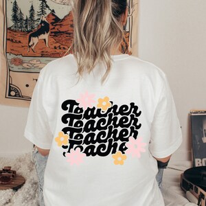 May include: White t-shirt with a black graphic that says "Teacher" four times in a retro font. The graphic is surrounded by pink and yellow flowers.