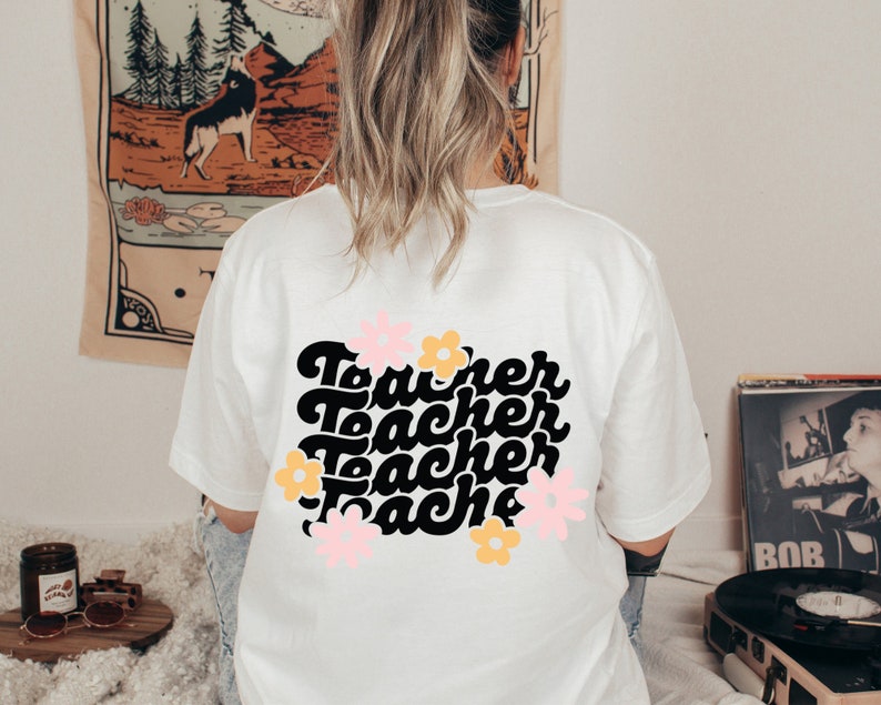 May include: White t-shirt with a black graphic that says "Teacher" four times in a retro font. The graphic is surrounded by pink and yellow flowers.