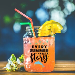 May include: A mason jar filled with a pink drink and a lemon wedge. The jar has a black and white sticker that says "Every Summer Has A Story" with a blue dolphin graphic.