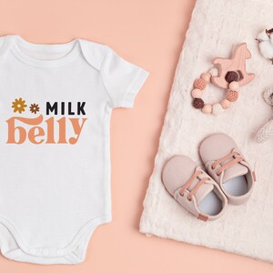 May include: White baby onesie with the text "* MILK belly" in orange and black. The onesie is on a pink background. There are baby shoes, a wooden toy, and a knitted toy on a white blanket.