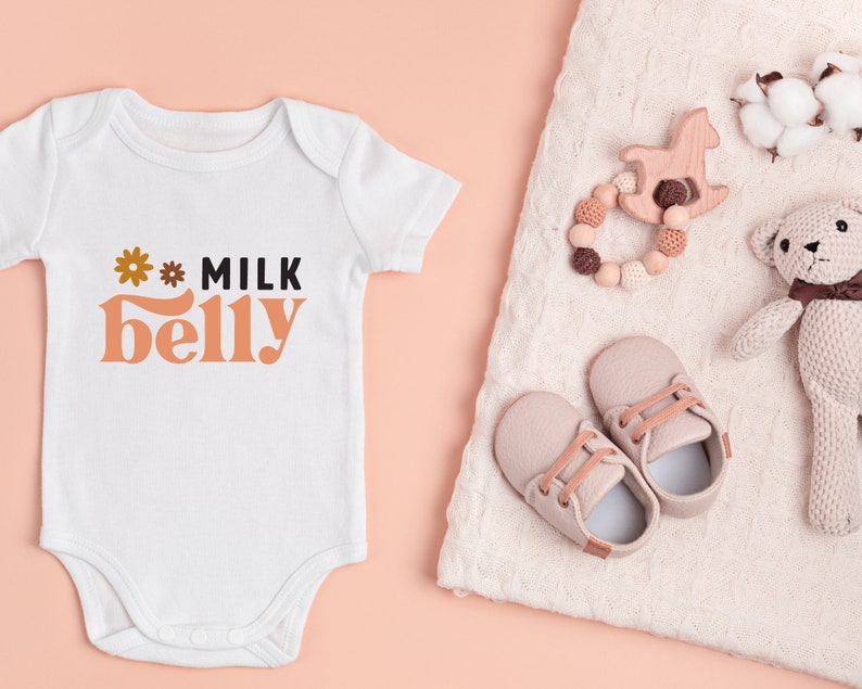 May include: White baby onesie with the text "* MILK belly" in orange and black. The onesie is on a pink background. There are baby shoes, a wooden toy, and a knitted toy on a white blanket.