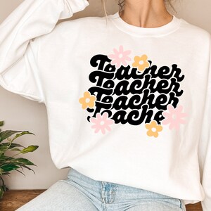 May include: White sweatshirt with a black graphic that says "Teacher" four times in a retro font. The graphic is surrounded by pink and yellow flowers.