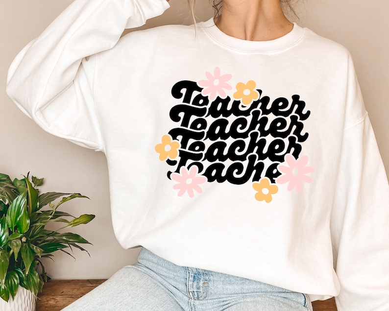 May include: White sweatshirt with a black graphic that says "Teacher" four times in a retro font. The graphic is surrounded by pink and yellow flowers.
